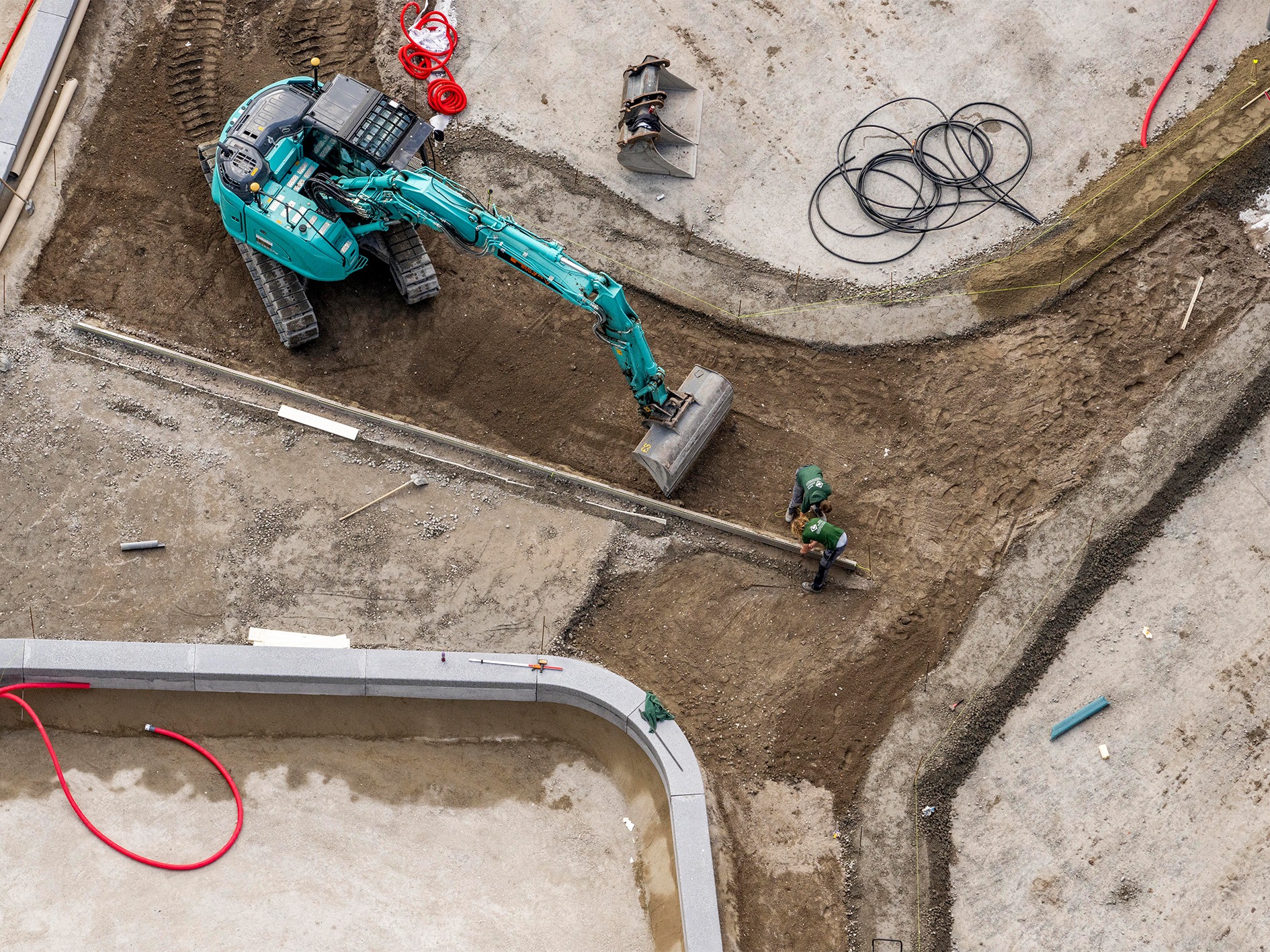 Luchtfoto van een bouwplaats waar een graafmachine en twee medewerkers werken aan de aanleg van een pad, omgeven door bouwmaterialen en kabels